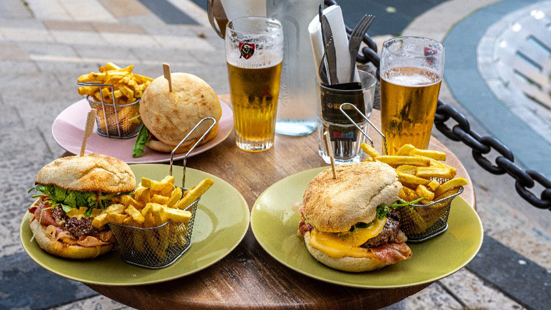 Trois burges frites dans des assiettes colorées. Les frites sont dans des petits paniers, on voit 2 bières